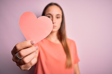 Young beautiful romantic woman holding paper heart shape over isolated pink background with a confident expression on smart face thinking serious