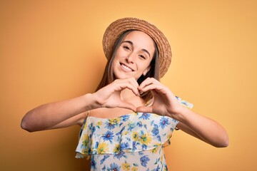 Young beautiful woman wearing casual t-shirt and summer hat over isolated yellow background smiling in love doing heart symbol shape with hands. Romantic concept.