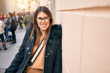 Young beautiful brunette woman wearing glasses smiling happy and confident. Standing with smile on face leaning on the wall at street of city