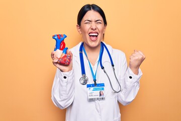 Young beautiful cardiologist woman wearing stethoscope holding heart over yellow background screaming proud, celebrating victory and success very excited with raised arm
