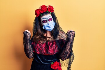 Young woman wearing day of the dead costume wearing medical mask screaming proud, celebrating victory and success very excited with raised arms