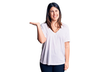 Obraz premium Young beautiful brunette woman wearing casual t-shirt smiling cheerful presenting and pointing with palm of hand looking at the camera.