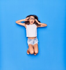 Adorable hispanic child girl wearing casual clothes and glasses surprised with open mouth. Jumping with hands on face  over isolated blue background