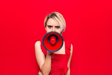 Young beautiful blonde woman with angry expression. Screaming loud using megaphone standing over isolated red background