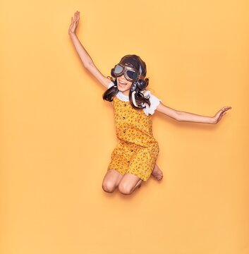 Adorable hispanic child girl wearing vintage aviator helmet and goggles smiling happy. Jumping with smile on face doing airplane gesture with arms open over isolated yellow background
