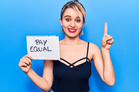 Young beautiful blonde woman holding pay equal paper smiling with an idea or question pointing finger with happy face, number one