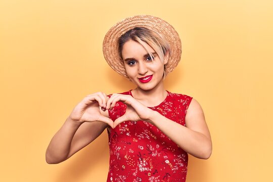 Young beautiful blonde woman wearing summer hat smiling in love showing heart symbol and shape with hands. romantic concept.
