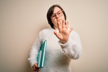 Young down syndrome student woman reading a book from library over isolated background with open hand doing stop sign with serious and confident expression, defense gesture