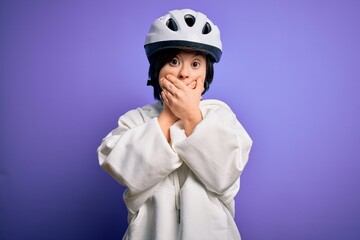 Young down syndrome cyclist woman wearing security bike helmet over purple background shocked covering mouth with hands for mistake. Secret concept.