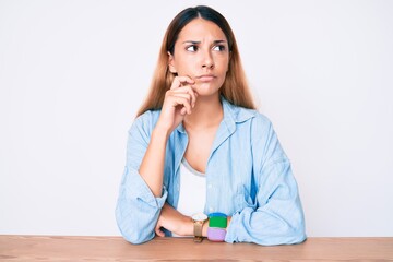 Young brunette woman sitting on the table wearing casual clothes serious face thinking about...