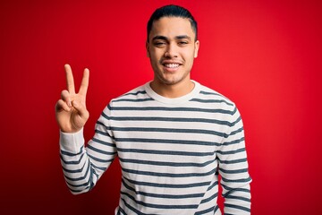 Young brazilian man wearing casual striped t-shirt standing over isolated red background smiling looking to the camera showing fingers doing victory sign. Number two.
