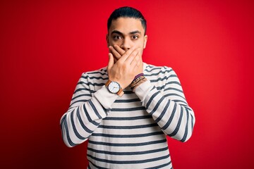 Young brazilian man wearing casual striped t-shirt standing over isolated red background shocked covering mouth with hands for mistake. Secret concept.