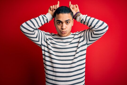 Young brazilian man wearing casual striped t-shirt standing over isolated red background doing funny gesture with finger over head as bull horns