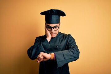 Young brazilian chef man wearing cooker uniform and hat over isolated yellow background Looking at the watch time worried, afraid of getting late