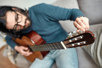 Fototapeta premium Close up of young caucasian man sitting on sofa at home and adjusting acoustic guitar. Main focus on a guitar. Playing musical instrument.