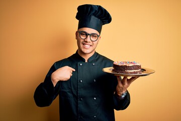 Young brazilian baker man wearing cooker uniform and hat holding tray with cake with surprise face...