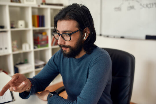 Close up of young bearded man teacher sitting at his workplace, looking at computer screen and giving online class through webcam at home