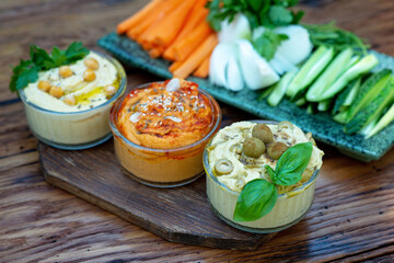 classic appetizer hummus with paprika and garlic, olives, chickpeas, tahini, olive oil, and oriental spices in a bowl on a wooden table with fresh vegetables. Mediterranean vegetarian snack.