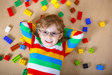 Little blond child with glasses playing with lots of colorful plastic blocks indoor. Kid boy wearing colorful shirt and having fun with building and creating