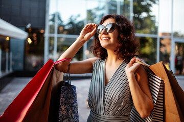 Happy girl stands near boutique shop holding a few bags with purchases. Young woman is glad to buy new clothes during sales season for a reasonable price. Shopping, discount, spend money concept.