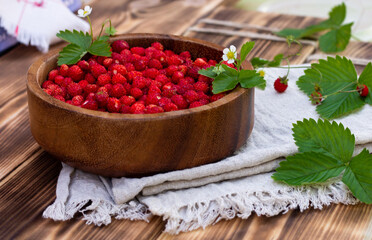 A wooden bowl of red ripe wild strawberries and flowers on old wooden surface.