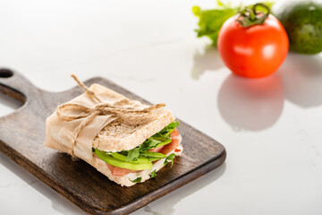 selective focus of fresh green sandwich with avocado and prosciutto on wooden cutting board on white marble surface