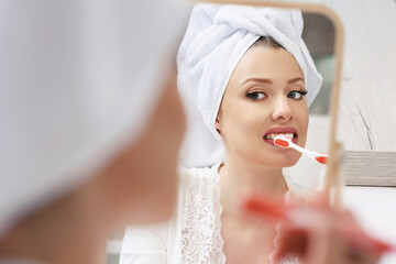 Young attractive woman with a bath towel on her head brushing her teeth in front of a mirror after a shower. Morning oral hygiene at home.