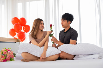 The girlfriend is glad to receive a rose from her boyfriend on the bed in the bedroom decorated with red heart balloons on Valentine's Day.