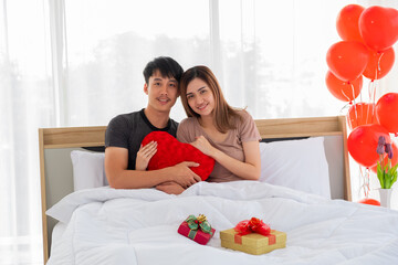 A young couple lovers hold a red heart shape pillow on their bedroom which have a red and a golden gift box on the bed.