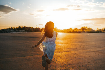 A little girl a child in a dress with long hair runs along an empty road in the sunset orange rays of the sun