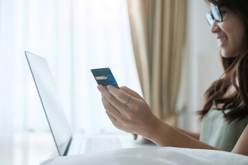 Happy woman holding credit card and using computer laptop for online shopping while making orders on bed in morning at home. Business, technology, ecommerce, digital banking and online payment concept