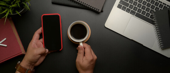 Male entrepreneur left hand using smartphone while right hand holding coffee cup on dark modern office desk