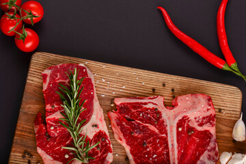 Fresh raw beef steak on wooden cutting board over black background with spices, top view.