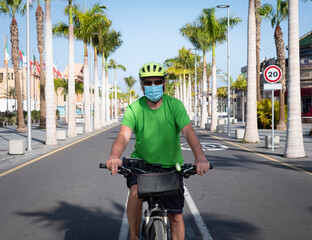 A senior man with bicycle and yellow helmet turning in deserted street wearing a face mask because of coronavirus crisis, no tourist in Tenerife island - active lifestyles in retired people