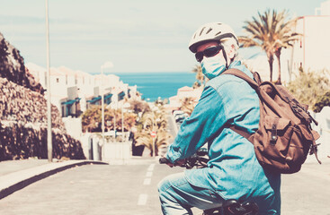 Senior man with bicycle and yellow helmet wearing face mask due to coronavirus infection, new normality, sea on background