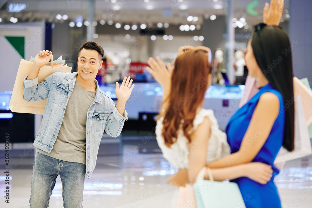 Poster Smiling handsome young Asian man waving with hand to his female friends in shopping mall