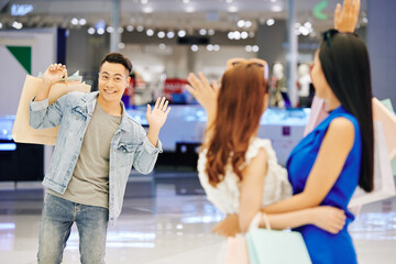 Smiling handsome young Asian man waving with hand to his female friends in shopping mall