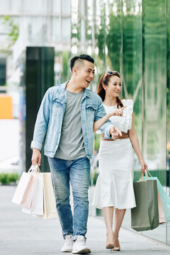 Happy young Asian couple with shopping bags walking in the street and discussing clothes in shop windows