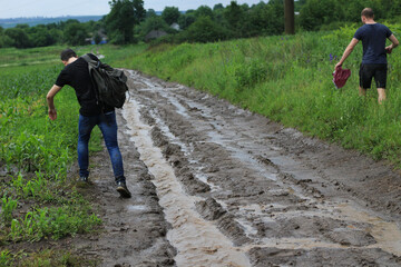 Tourists outside the city fell in heavy rain and walk along a soggy dirt road towards civilization - obrazy, fototapety, plakaty