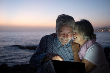 Cheerful senior couple using digit tablet sitting outdoor at night. Palm tree and night lights on background - concept of happy retired people tech and social