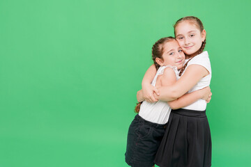 Two little sisters in school uniform hugging each other, having fun on a green background