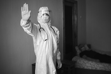 Black and white image of doctor in PPE suit, face mask, gloves, phonendoscope, visiting patient at home during covid-19 quarantine, showing stop sign, lying patient on background
