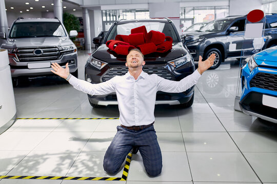 Lucky Owner. Excited Young Caucasian Man Screaming Happily With His Arms Raised Standing On His Knees Near His New Car At The Car Dealership