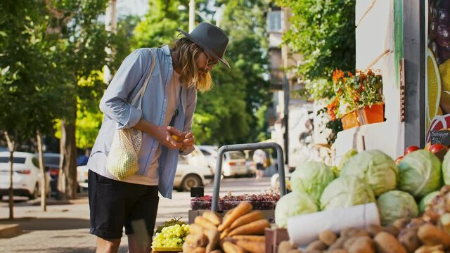 Young male with reusable mesh shopping bag is choosing apricots or plums at small street market with vegetables and fruits. Zero waste, eco friendly
