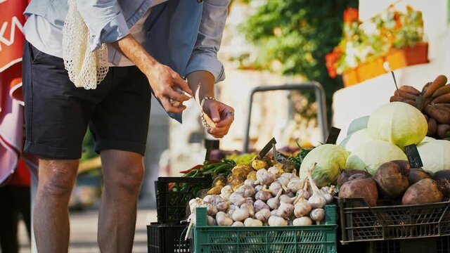 Unrecognizable fellow in casual outfit, mesh bag. Choosing garlic at small street market with vegetables and fruits. Zero waste. Slow motion, close up