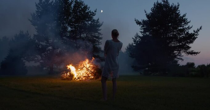 Natural woman wearing dress looking at bonfire in garden at midsummer night