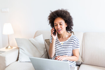 Happy woman sitting on sofa with laptop and talking on phone at home. Young successful businesswoman working from home while talking at phone. College student studying on laptop and using phone.