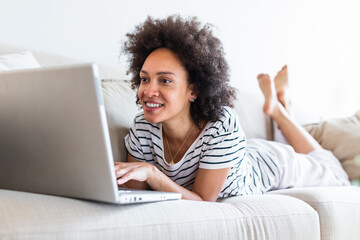 Young beautiful woman with curly hair working on laptop computer while sitting on the sofa at home, heck on oline shops for cyber monday sales. Technology woman concept alternative office freelance