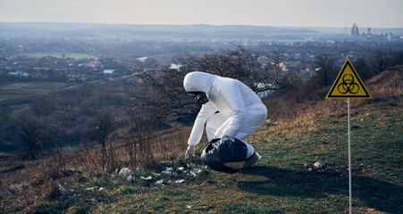 Environmentalist in protective suit and gas mask picking up garbage in abandoned grassy field, with biohazard sign. Concept of ecology, environmental pollution and biological hazard.