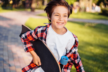 Happy young guy running along the alley on a sunny summer evening. Holding his skateboard in hands. Actively spending summer holidays. Dressed in a plaid shirt and white T-shirt